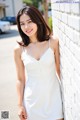 A woman in a white dress leaning against a brick wall.